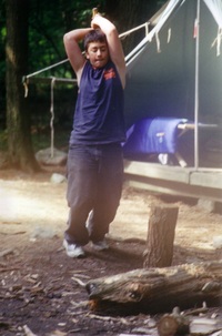 A scout is holding an ave over his head, looking at a log on the ground that is standing up vertically.