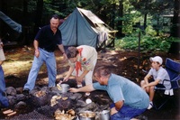 Four people are staring at a fire pit.  Two of them are holding chickens over the fire pit.