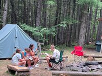 Three scouts are sitting on a wooden log bench, and other is facing them in a chair, reading out of a book.