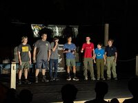 Seven scouts stand on a stage in front of people at night.