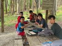 Six scouts sitting around a picnic table.  A chess board is in the middle, and other scouts are playing cards.