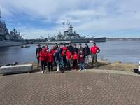 The troop stands on the shore.  Behind them are several battle ships.