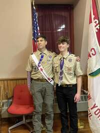 Two people in class A uniforms smile for the camera in front of the American and Troop flags.