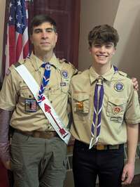 Two people in class A uniforms smile for the camera in front of the American Flag.