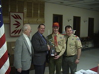 Four people smiling for the camera.  The two folks on the right are in Class A uniforms.  The two people in the middle are shaking hands with each other.