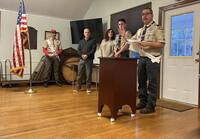 Five people stand in front of a podium.  Three are in a class A uniform.  One is speaking behind the podium.