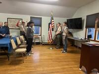 Two people standing in front of the room in class A uniforms.  One is holding an America Flag, the other is giving the scout sign.