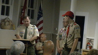 Two people standing in Class A uniforms.  The person on the left if at a podium, reading from something on it.