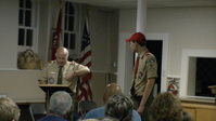 Two people standing in Class A uniforms.  The person on the left if at a podium, reading from something on it.