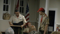 Two people standing in Class A uniforms.  The person on the left if at a podium, reading from a folder.