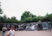 A man in a white Troop 53 class B tee-shirt facing the camera smiling.  In the background, scouts sit looking at waterfalls.