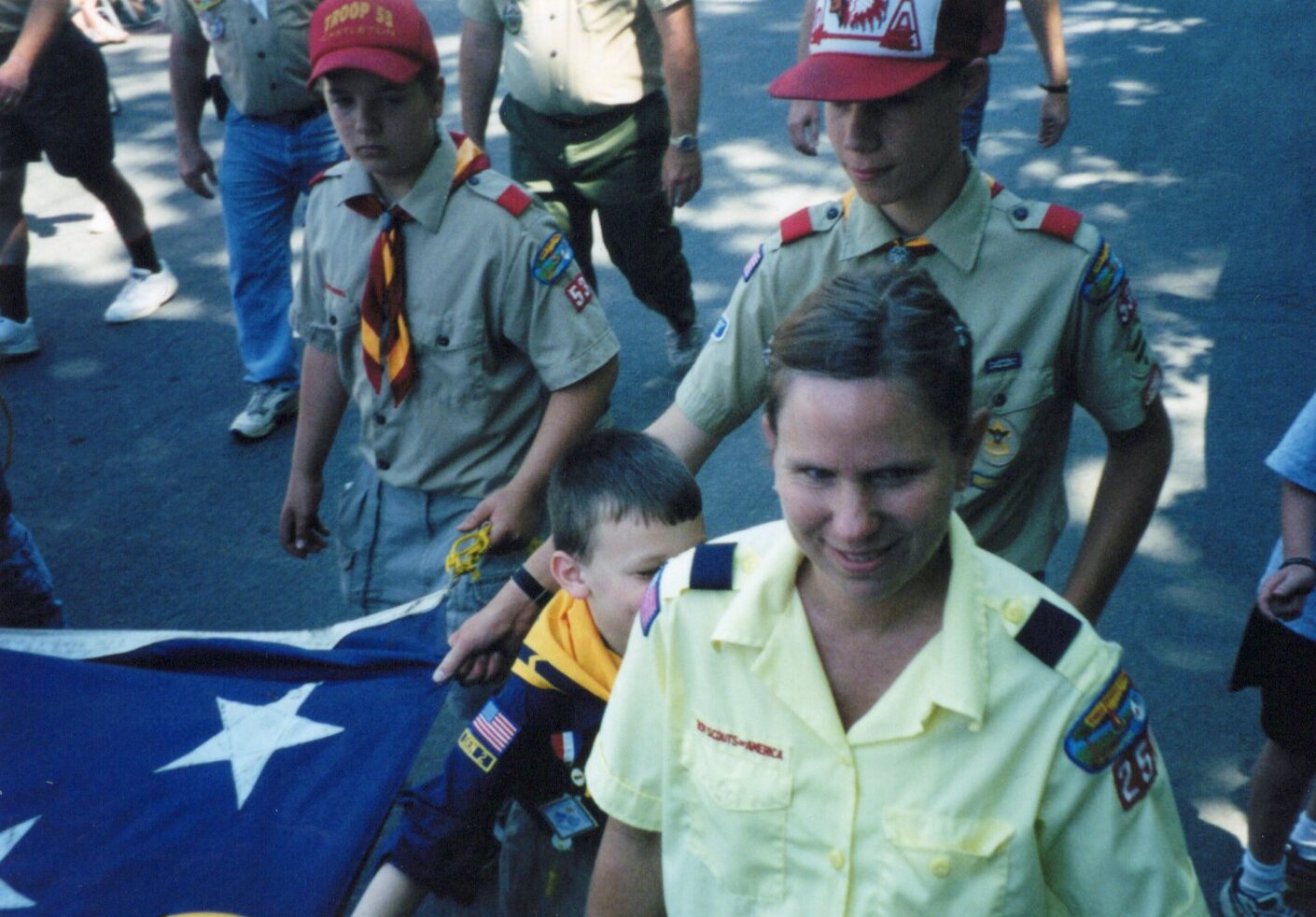 Four people holding up the giant America flag from the back corner.  All are in uniform.  One is an adult woman, the rest are boys.