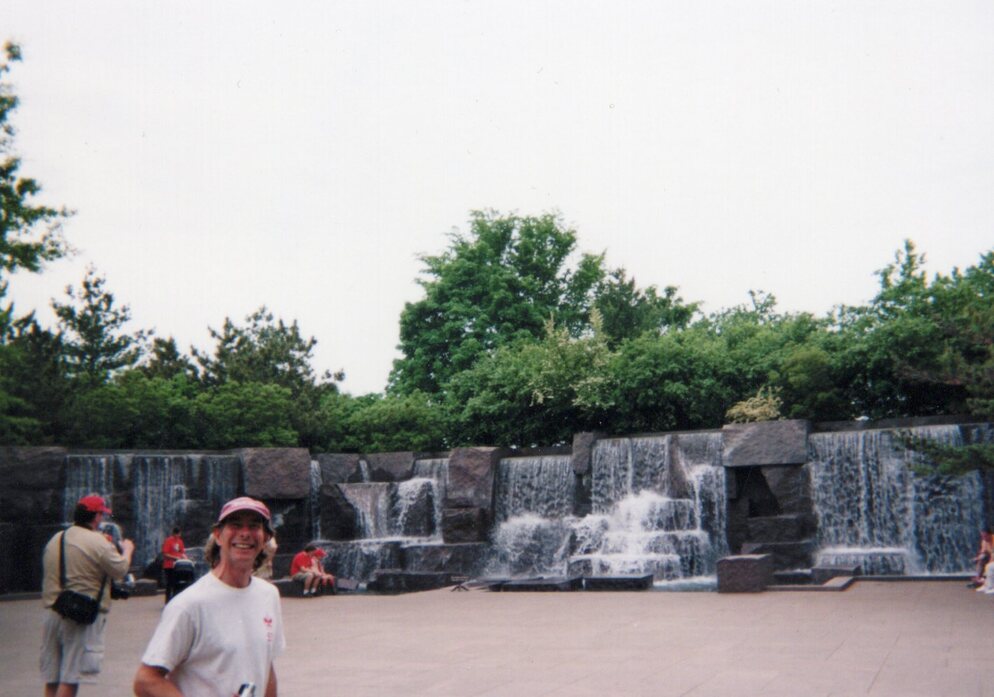 A man in a white Troop 53 class B tee-shirt facing the camera smiling.  In the background, scouts sit looking at waterfalls.