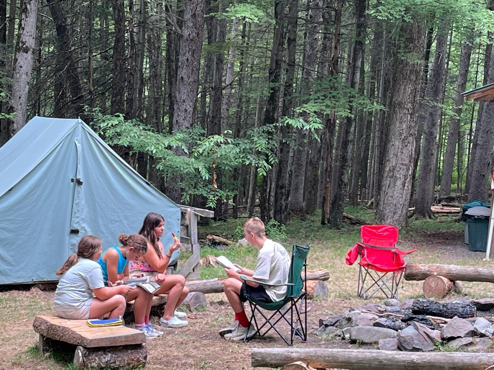 Three scouts are sitting on a wooden log bench, and other is facing them in a chair, reading out of a book.