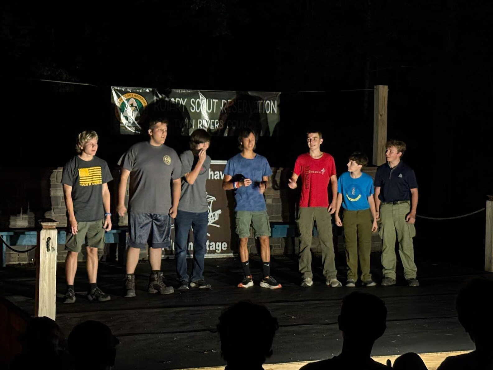 Seven scouts stand on a stage in front of people at night.