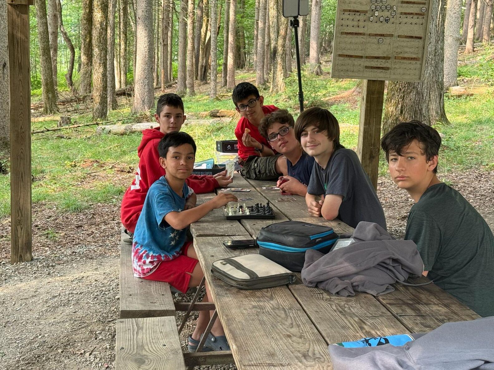 Six scouts sitting around a picnic table.  A chess board is in the middle, and other scouts are playing cards.