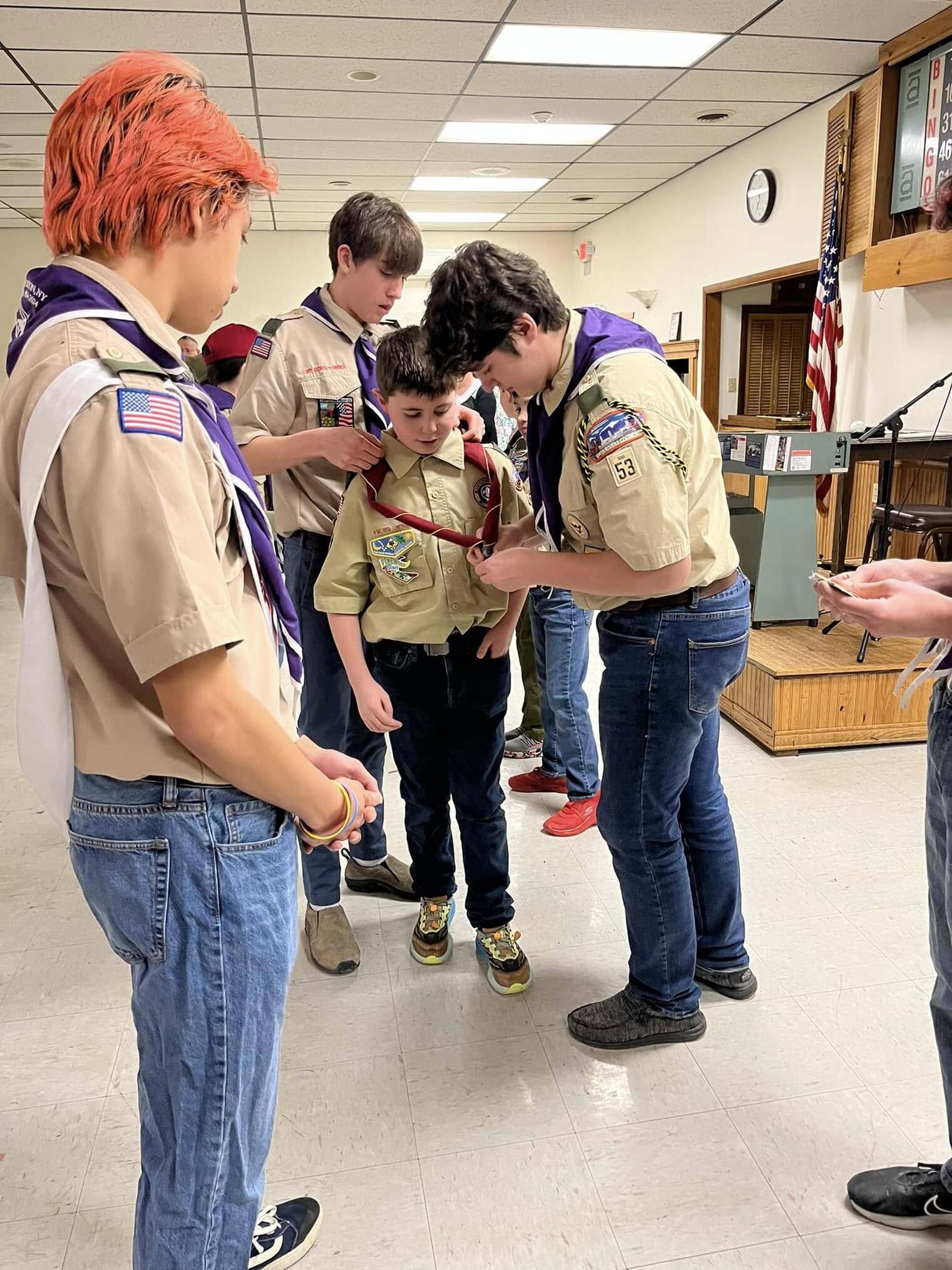 Two older scouts help a younger cub scout put on the Scouts BSA neckerchief.  A scout with red-dyed hair looks towards them.