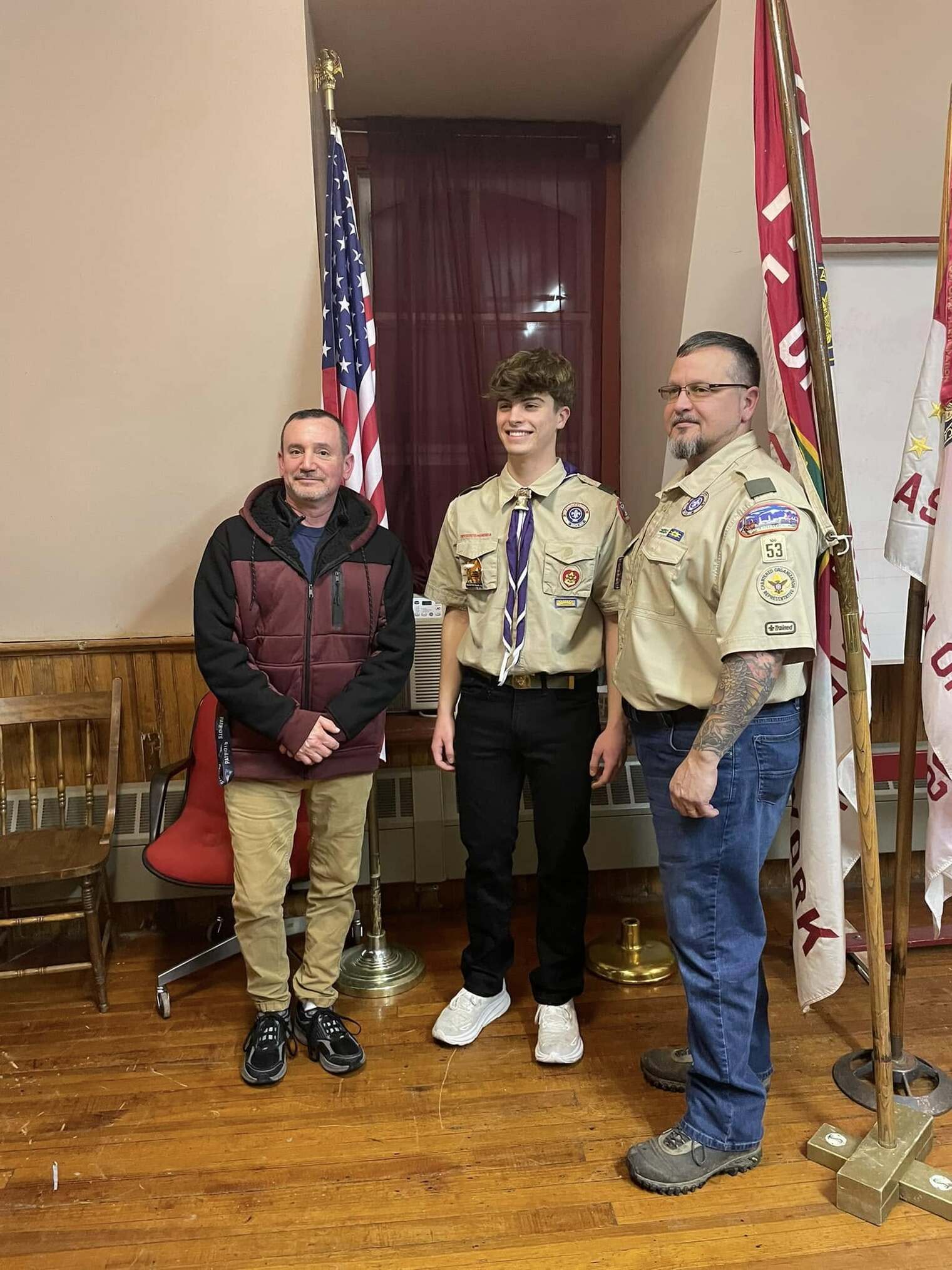 Three people stand in front of the American flag and troop flags.  The two on the right are in class A uniforms.  Ben is in the middle.