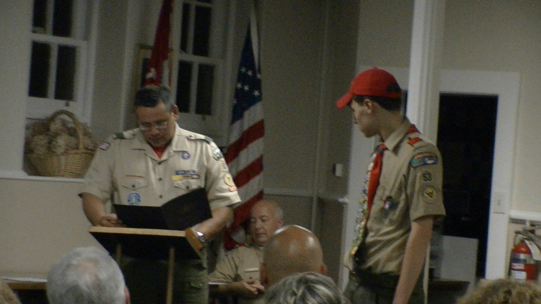 Two people standing in Class A uniforms.  The person on the left if at a podium, reading from a folder.