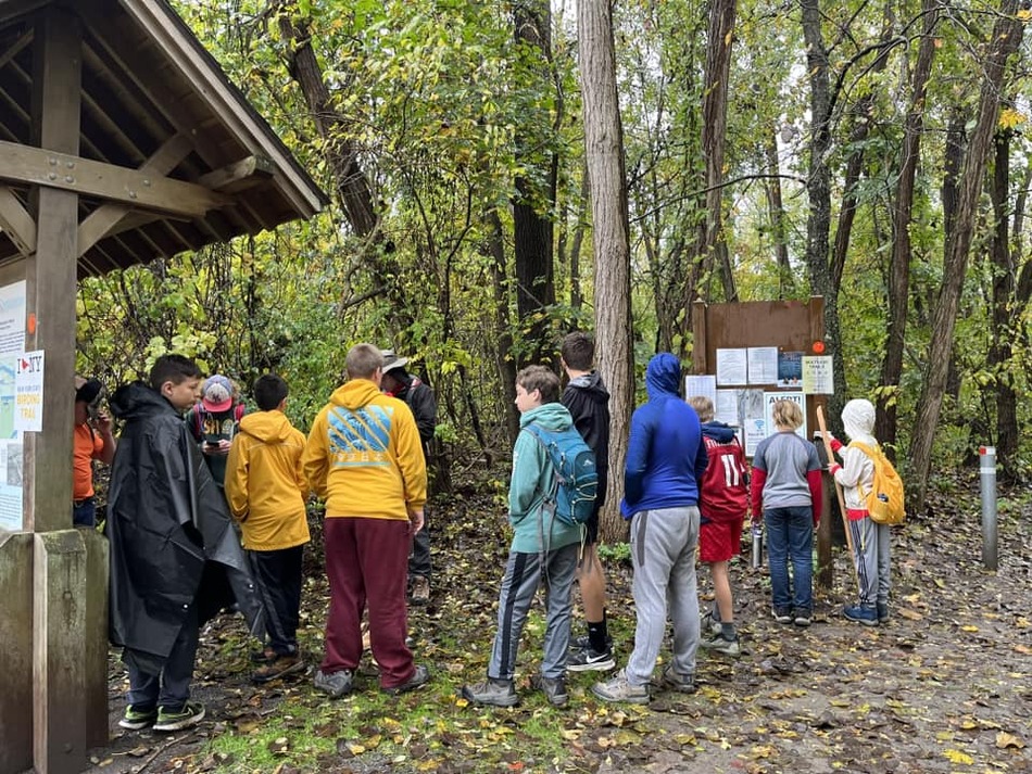 A group of scouts gathered by a trail head looking at a sign with a lot of notices on it.